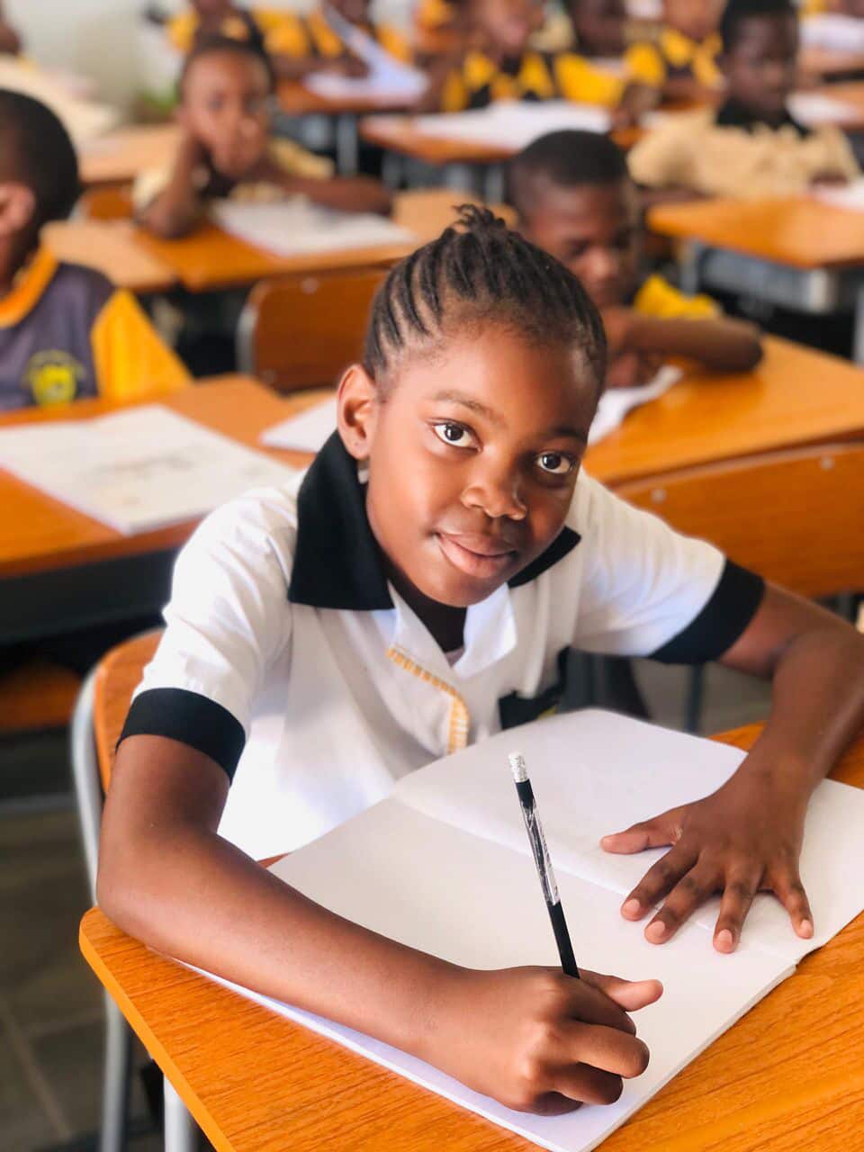 Learner smiling at her desk with an open exercise book and pencil in a full classroom