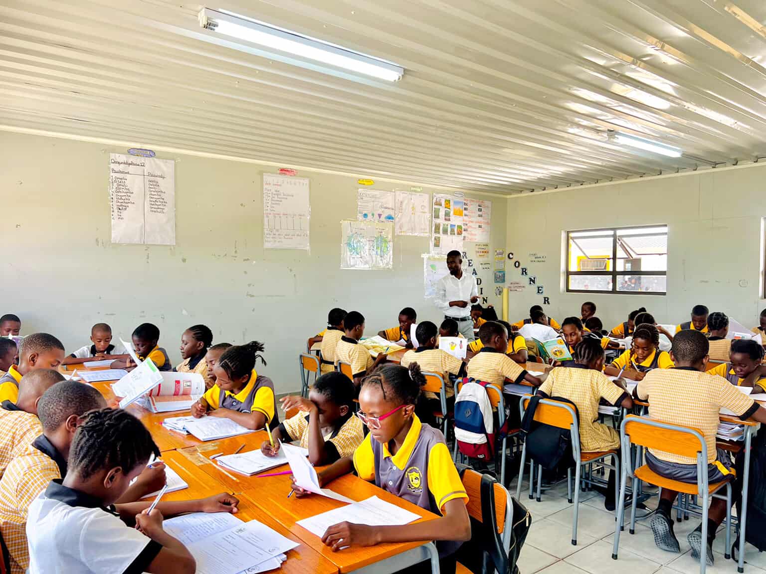 Wide view of a large classroom with learners reading and writing while a teacher leads the lesson