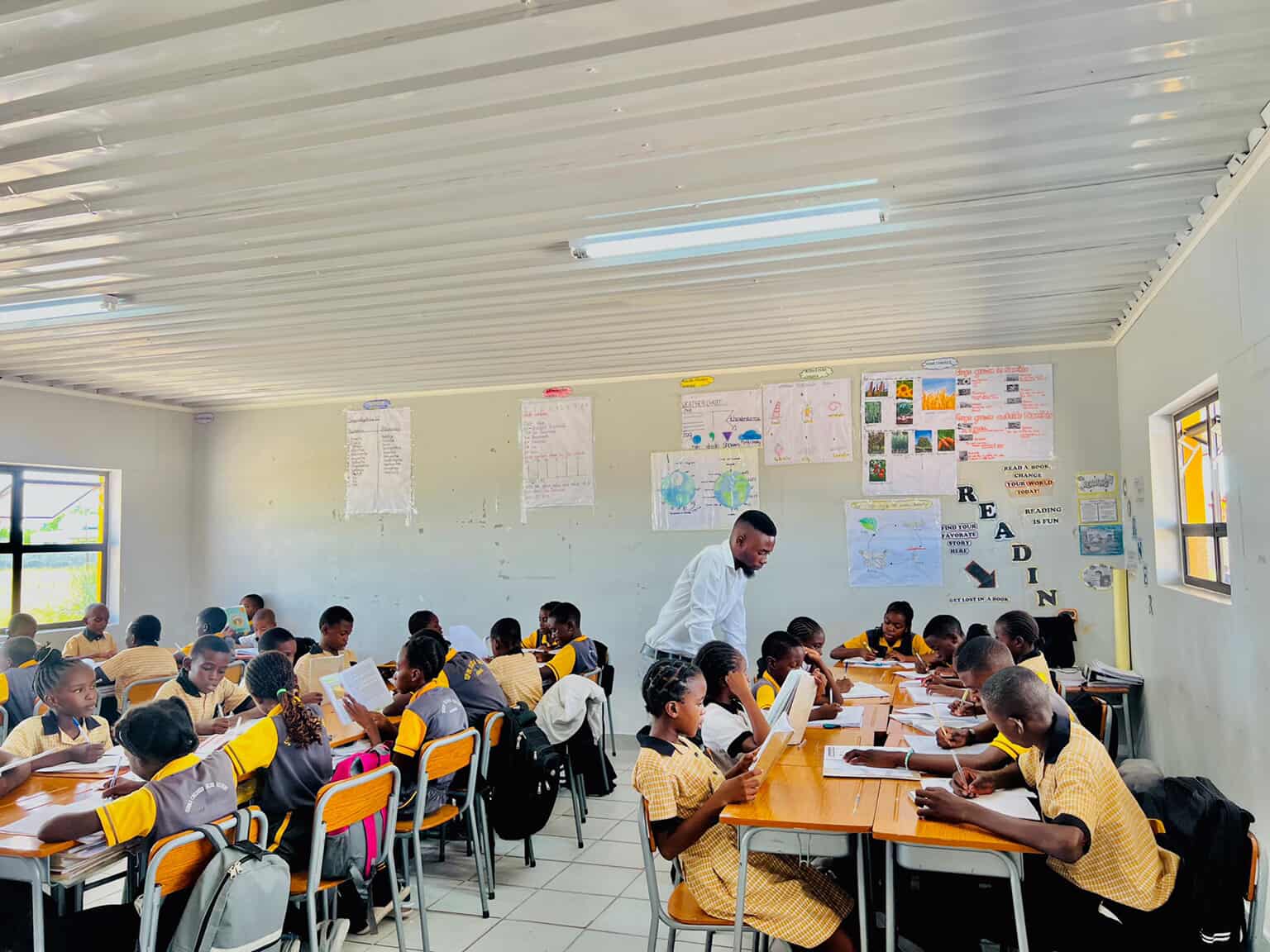 Teacher walking between desks in a full classroom while learners read and write
