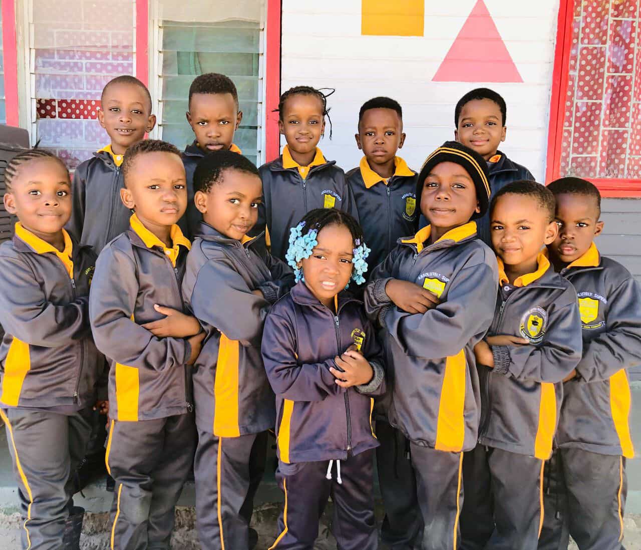 Group of preschool learners in grey and gold tracksuits posing together outside their classroom
