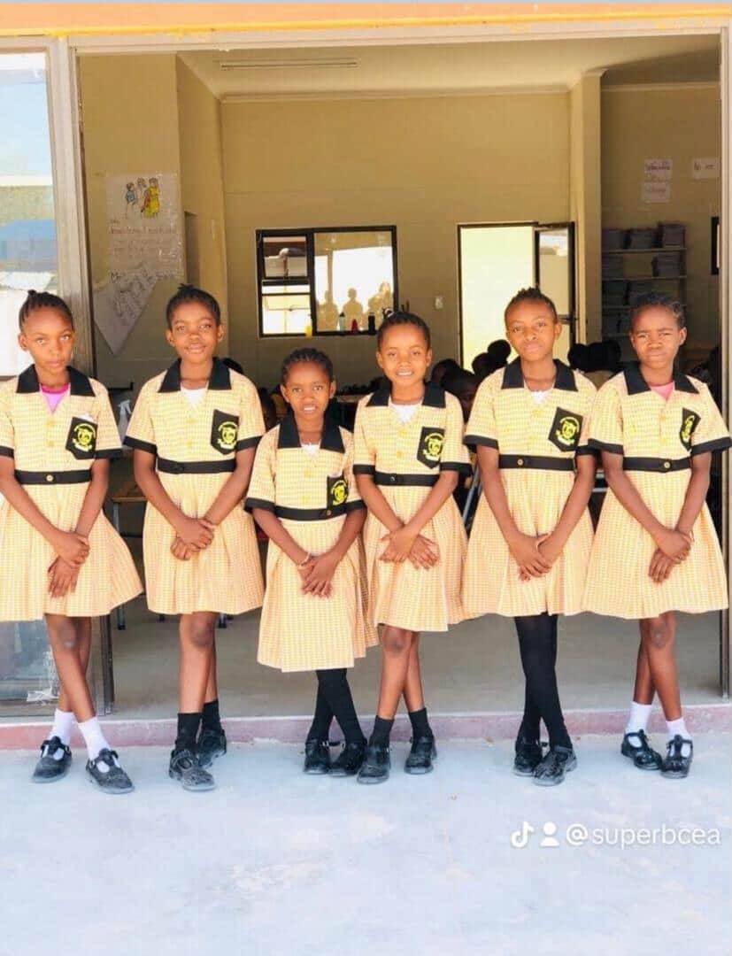 Six learners in checked school dresses standing outside their classroom