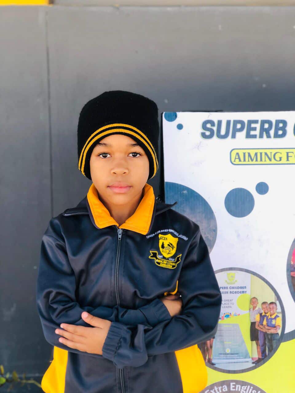 Learner in a Superb Children English Academy jacket and beanie standing by a school banner