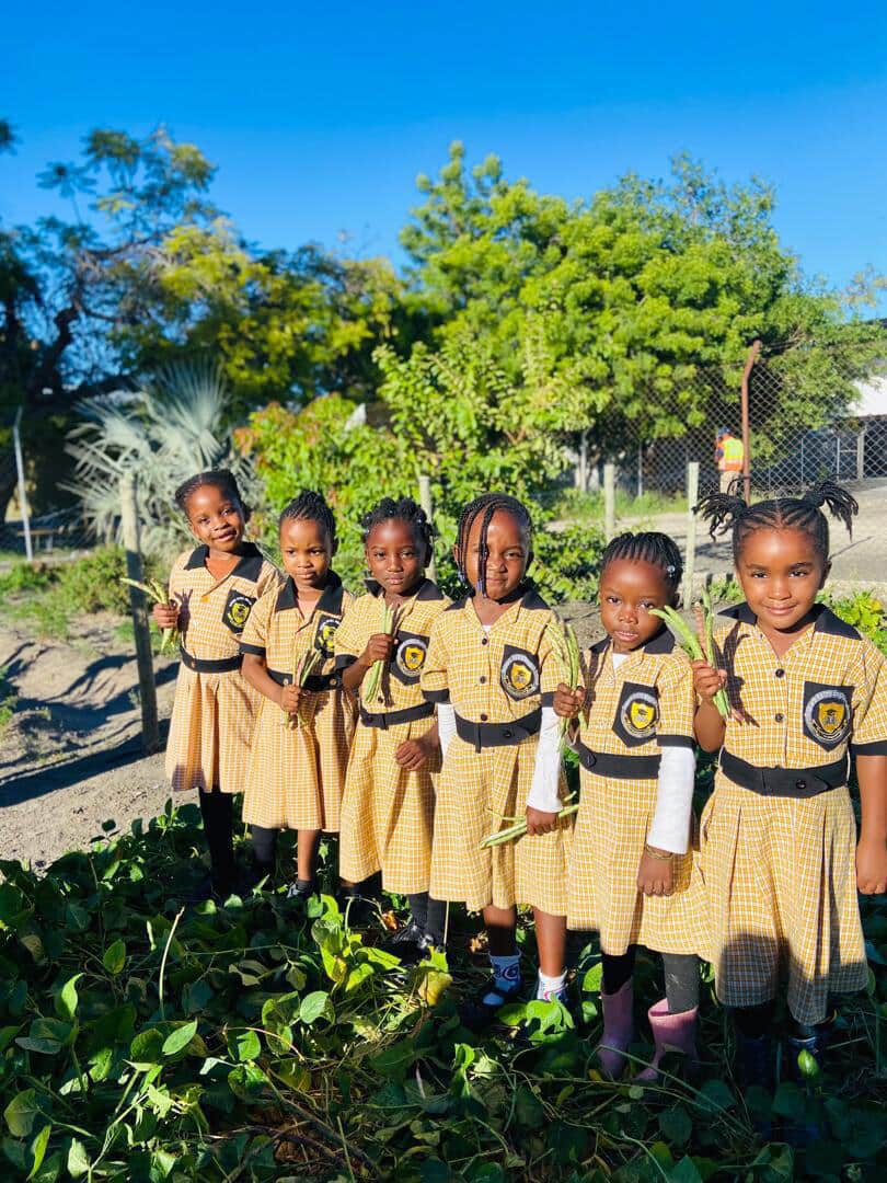 Six preschool learners in checked uniforms holding freshly picked beans in the school garden