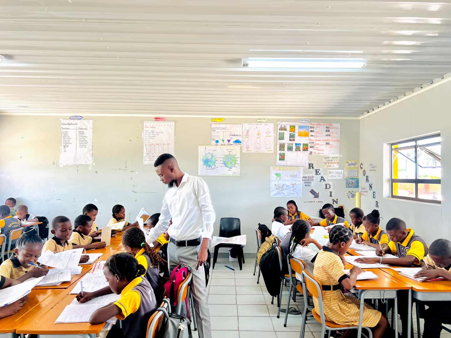 Teacher handing out worksheets to learners in a bright classroom with a reading corner